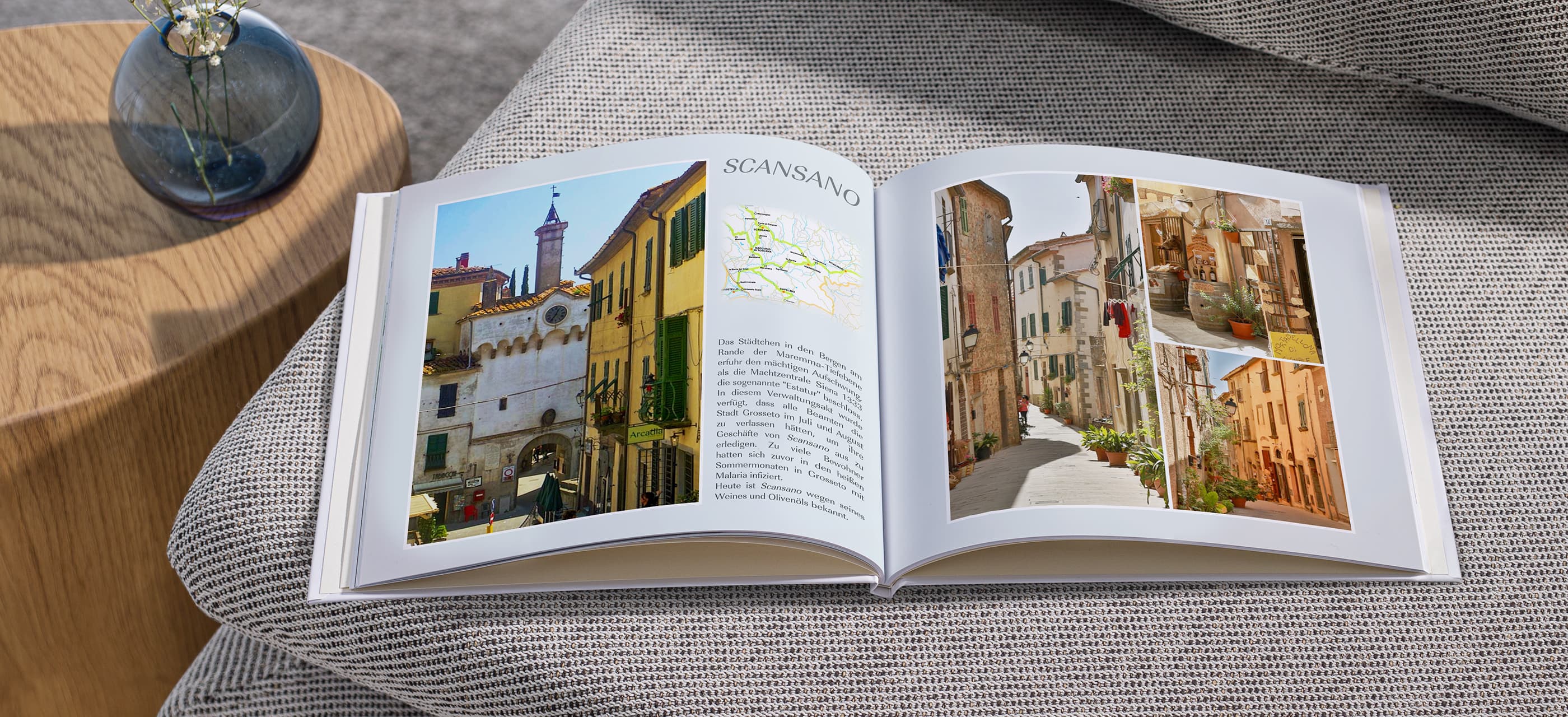 Ein geöffnetes Fotobuch aus dem Italienurlaub liegt geöffnet auf einem hellgrauen Sofa, dass die Häuser und Straßen des Ortes Scansano zeigt, daneben ein kleiner Couchtisch mit einer dunklen Blumenvase aus Glas.